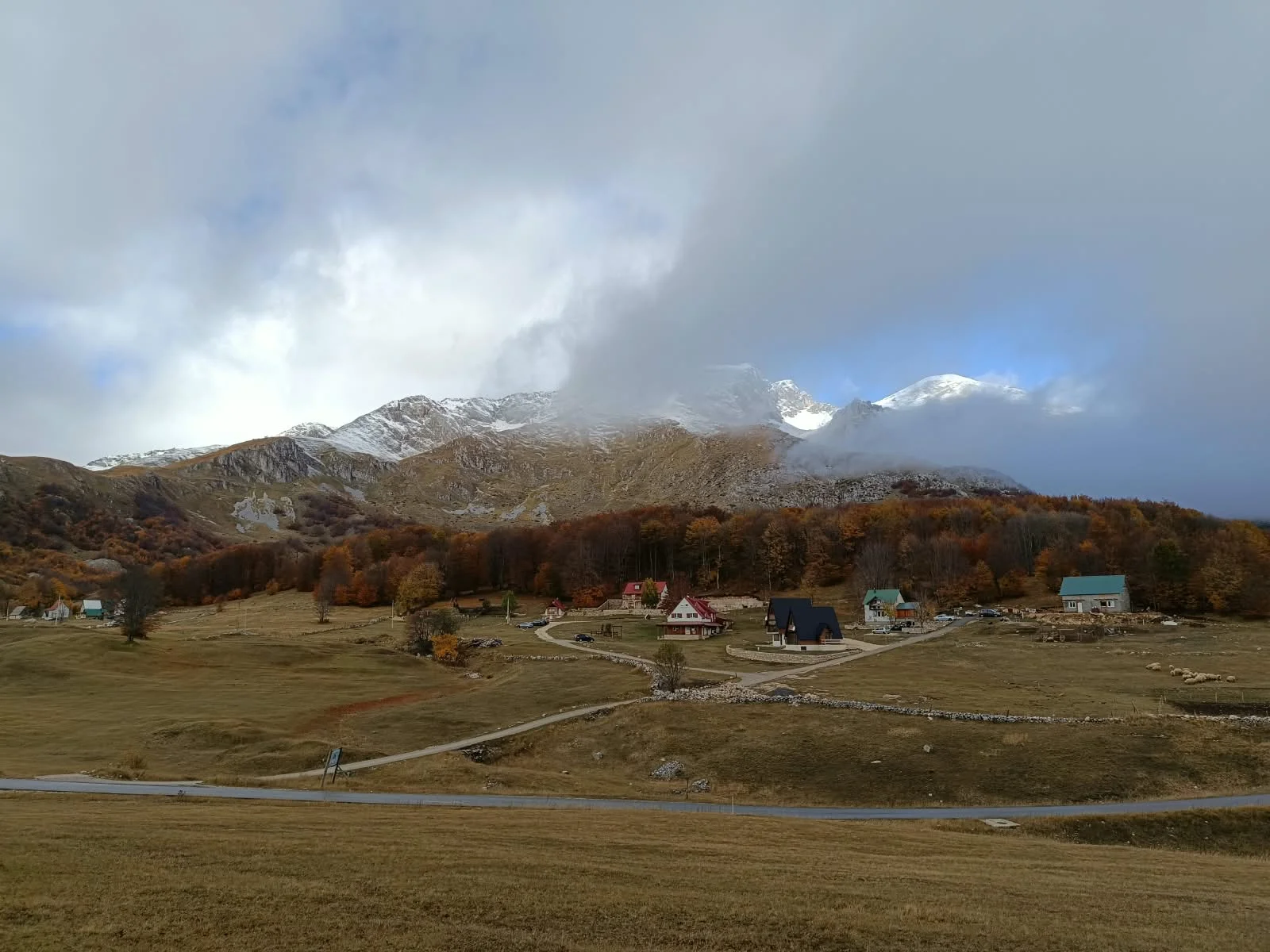 Autumn valley in Durmitor: houses by a beech forest and snowy peaks in the clouds